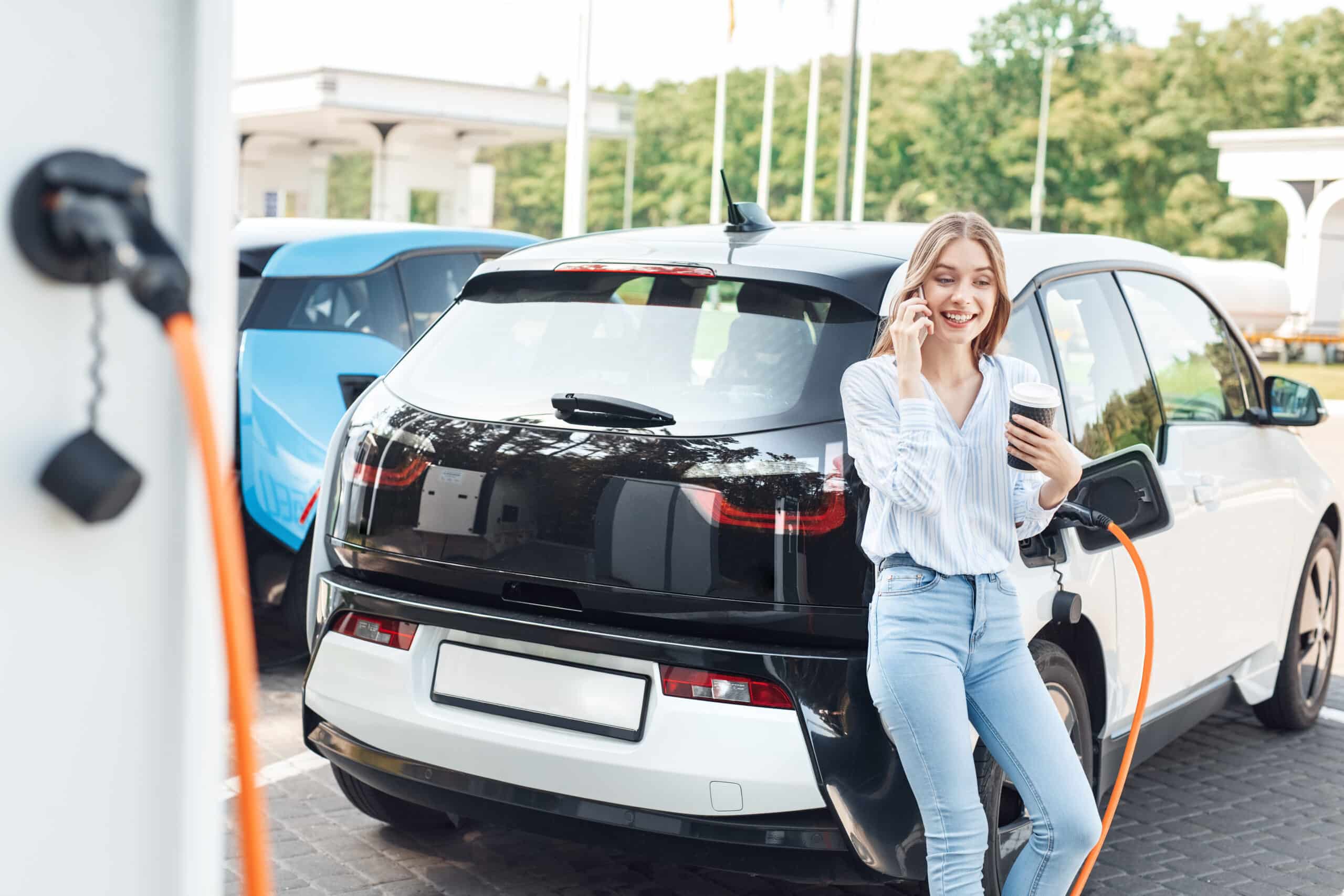 Young adult woman standing on parking, charging electric car, talking on smartphone